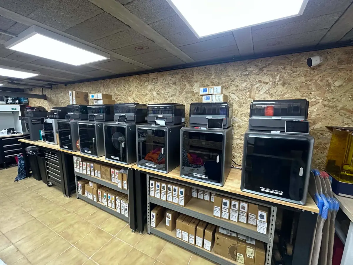 Rangée d'imprimantes Anycubic Kobra S1 alignées sur un long établi, murs en OSB et racks de filament en dessous