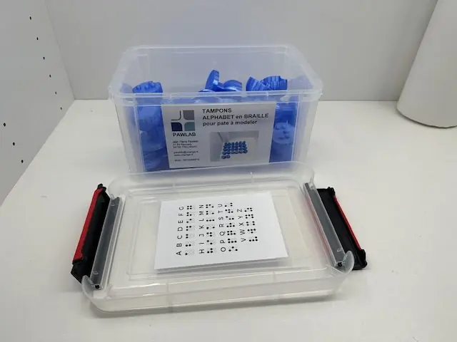 Boîte transparente remplie de tampons braille bleus, étiquetée Pawlab, avec sa planche alphabet
