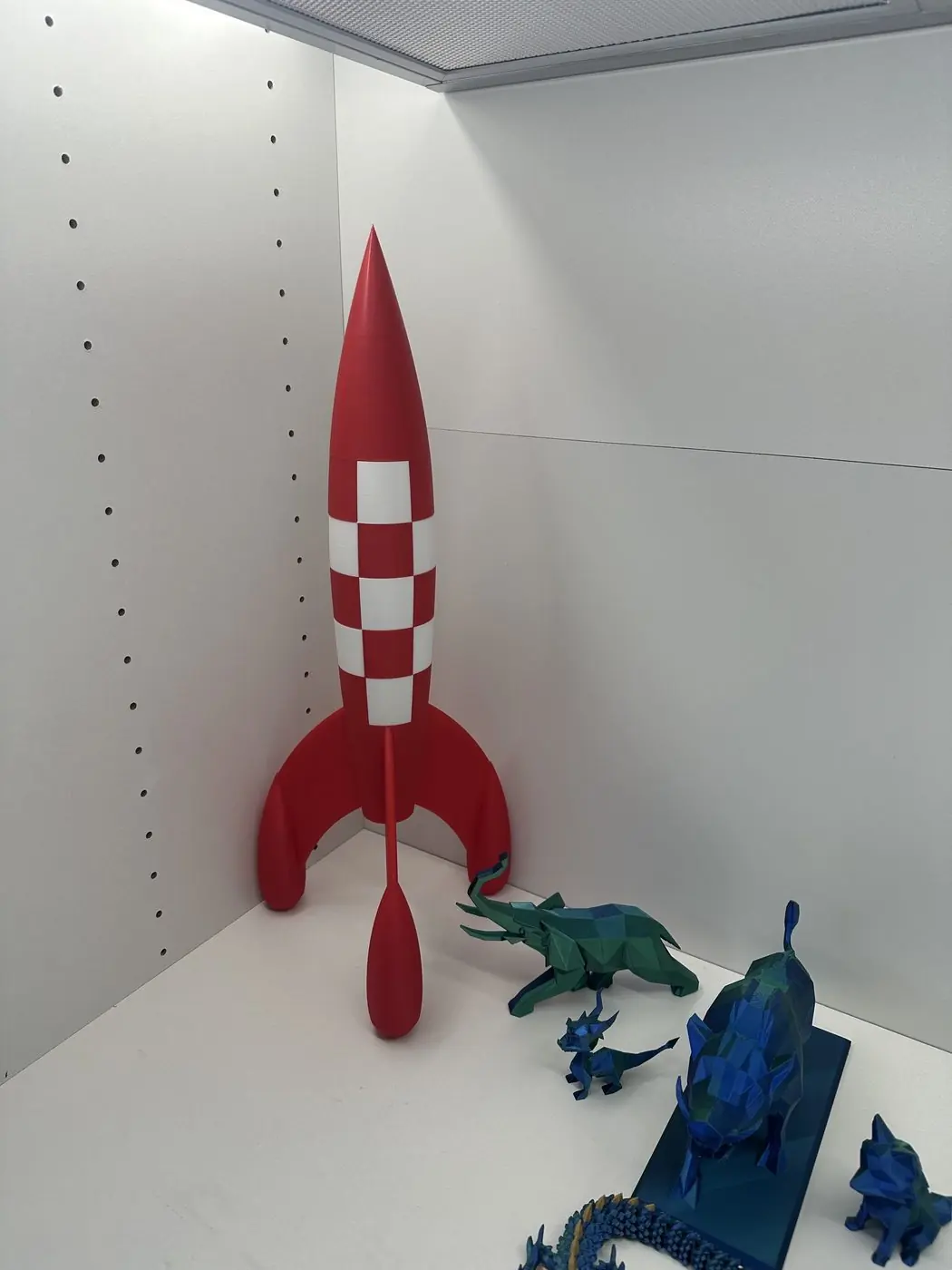 Grande fusée décorative rouge et blanche imprimée en 3D, posée sur une étagère à côté de figurines low-poly